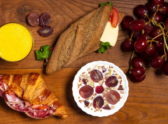 Flat lay of croissant, baguette, grapes, yogurt, and juice on a wooden table.