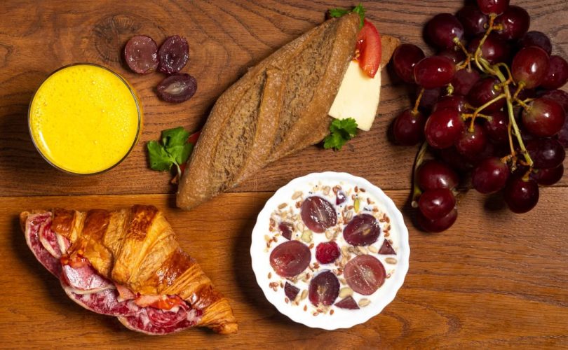 Flat lay of croissant, baguette, grapes, yogurt, and juice on a wooden table.