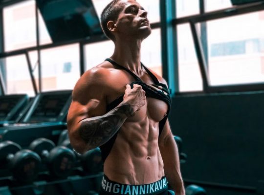 Muscular man showing abs and tattoos in a gym setting.