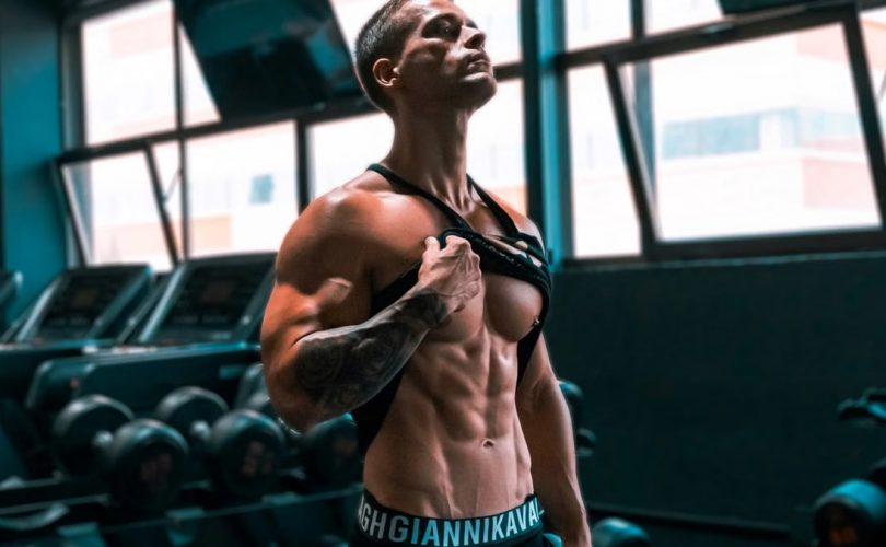 Muscular man showing abs and tattoos in a gym setting.